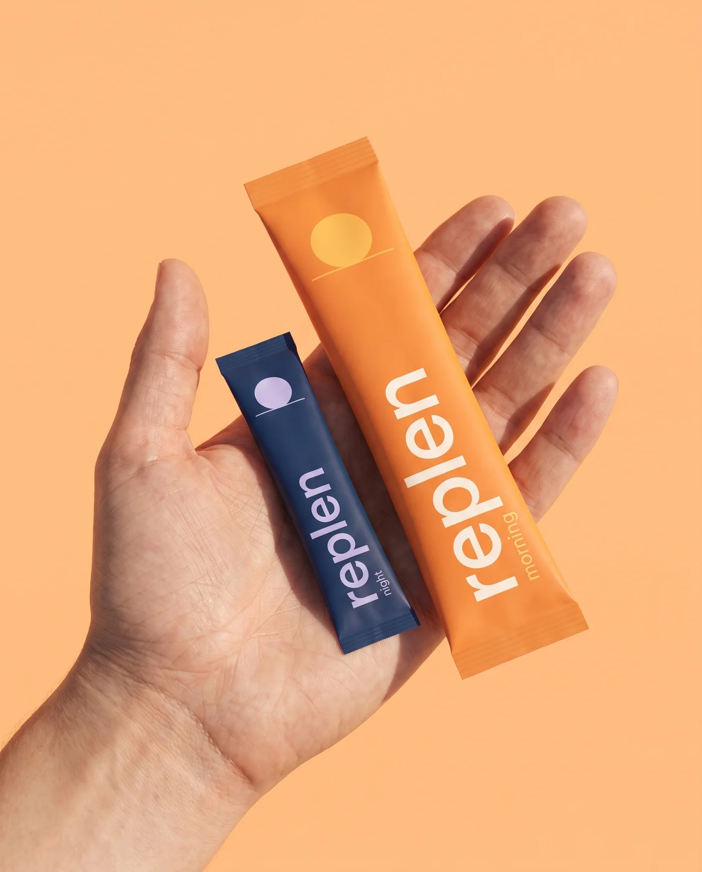An open hand holding both Replen sticks: the larger orange morning stick and the shorter midnight-blue night stick
