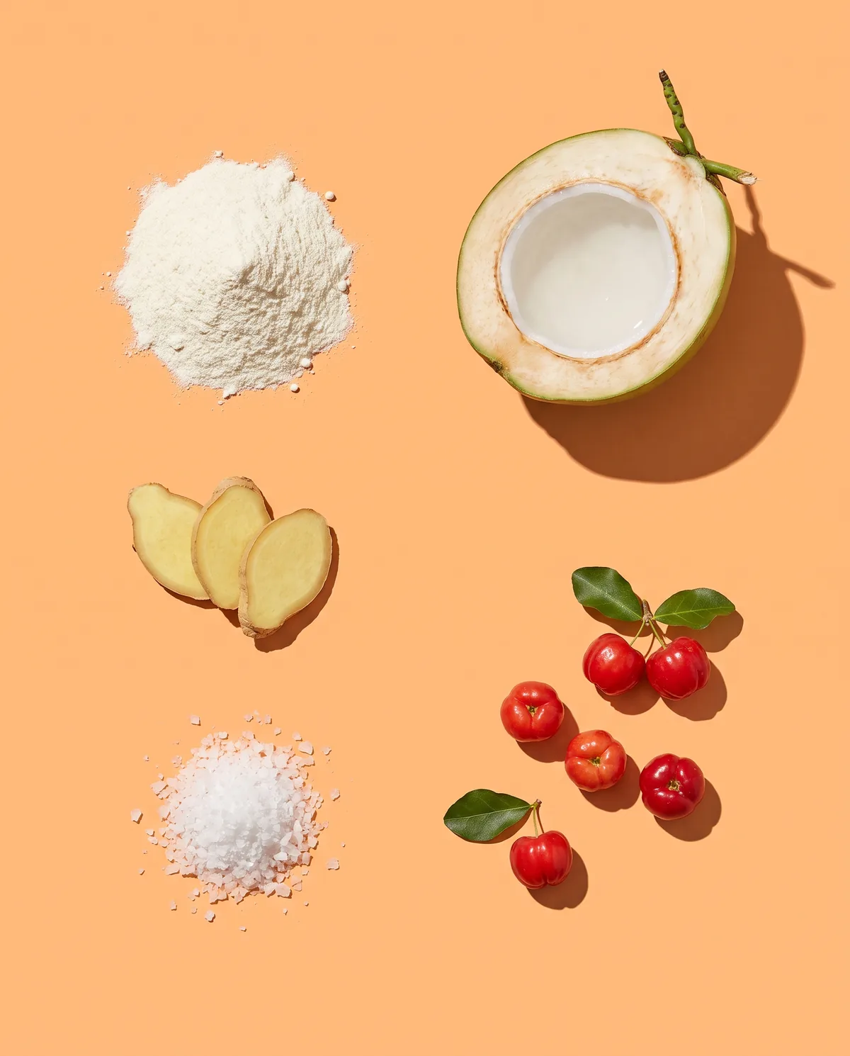Whey powder, a halved young coconut, ginger slices, sea salt and acerola cherries laid out on an apricot backdrop