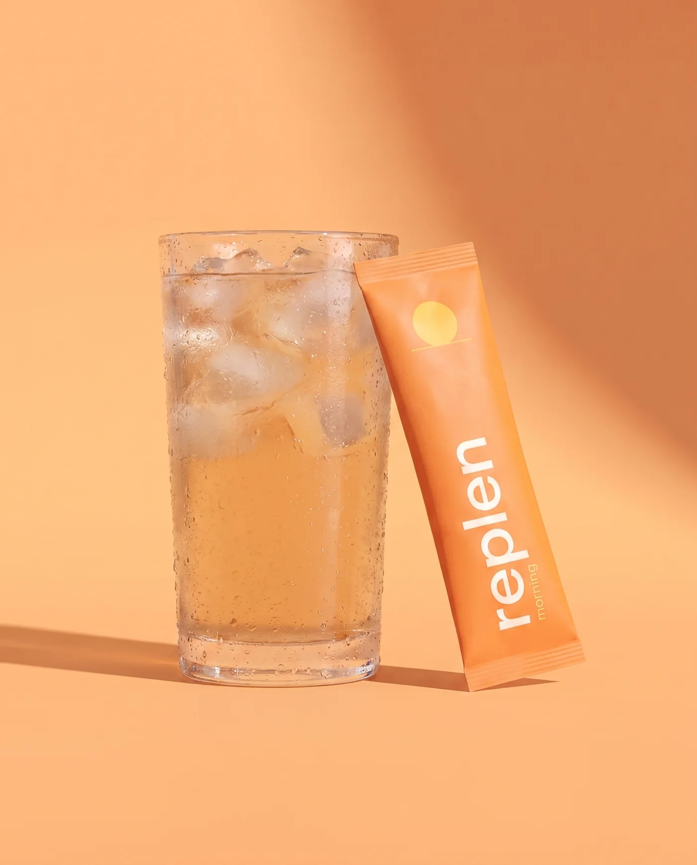 A tall glass of pale apricot drink with ice, the orange morning pack leaning against it