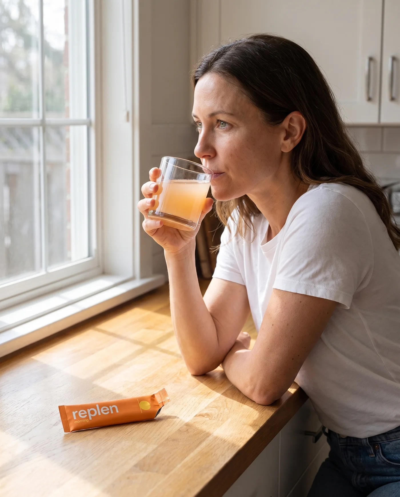 A woman at a bright kitchen counter taking a small sip from a short glass, the morning pack lying in front of her