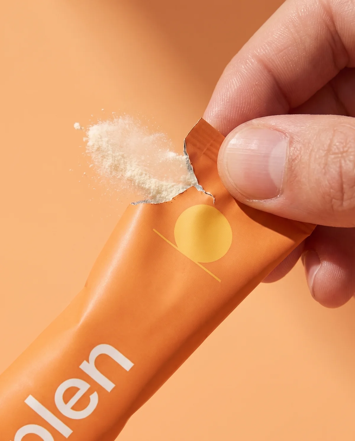 Two fingertips tearing open the orange morning pack, a puff of powder escaping